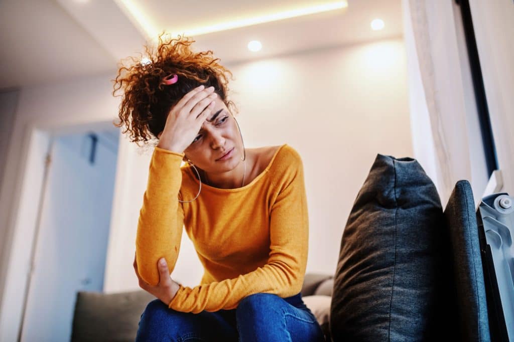 Woman in yellow sweater sitting on couch, holding her head in distress, representing anxiety and mental health challenges.