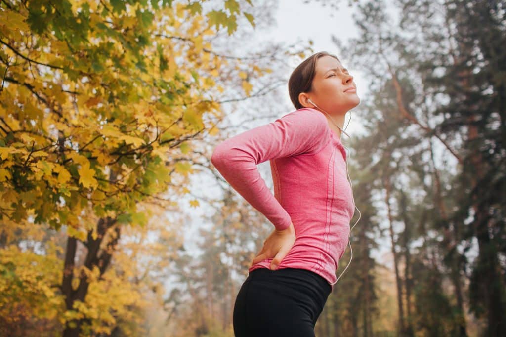 Woman in a pink long-sleeve shirt experiencing back pain outdoors, surrounded by autumn foliage, illustrating the impact of chronic pain and natural remedies like CBD and THC.