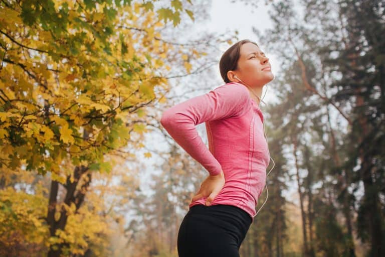 Woman in a pink long-sleeve shirt experiencing lower back pain outdoors among autumn foliage, illustrating the topic of using CBD and THC for pain relief.