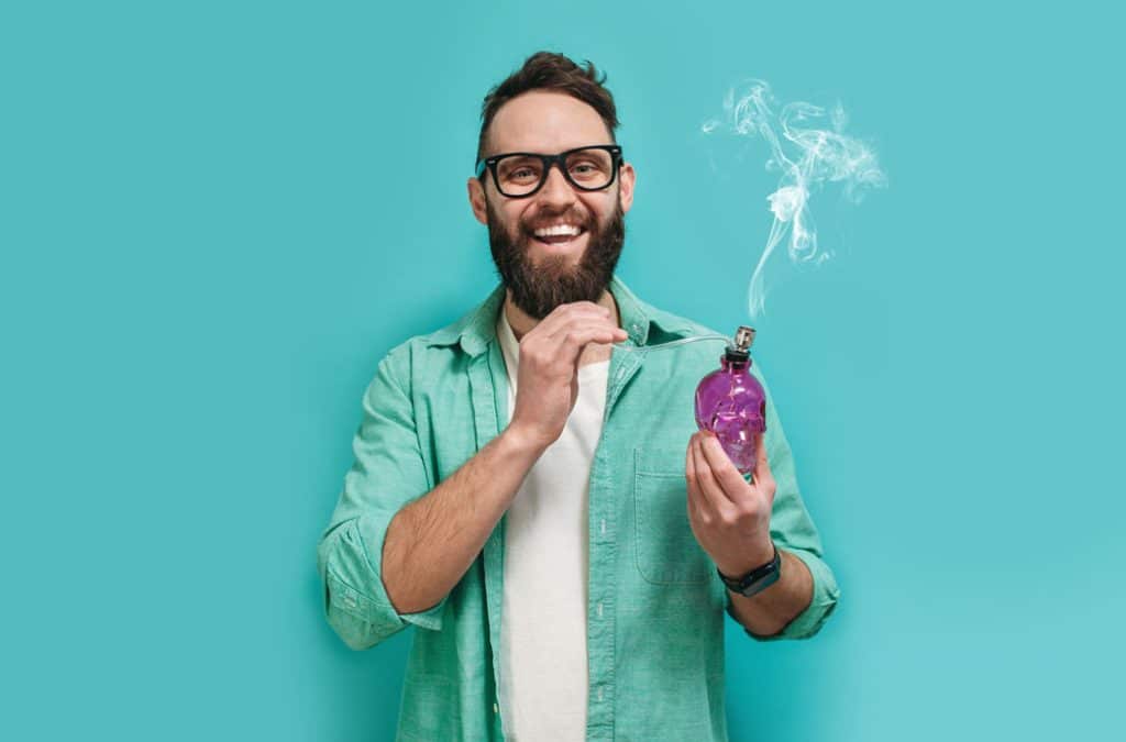 Man smiling and holding a purple smoking device with smoke, against a turquoise background, representing recreational use of cannabis products.