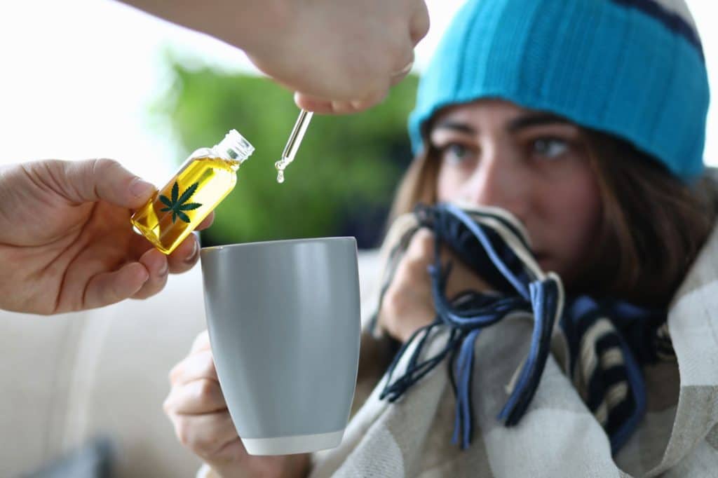 Person in a blue beanie holding a blanket, while a hand pours CBD oil from a dropper into a gray mug, illustrating the use of cannabis for stress relief and anxiety management.
